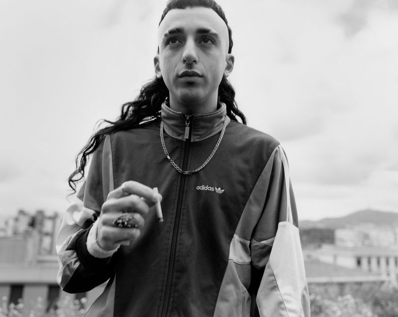 Monochrome portrait of a man with long dark curly hair wearing a zip-up track jacket holding a cigarette against an outdoor rooftop backdrop