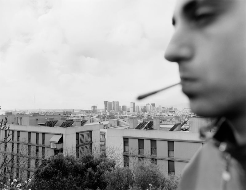 Close-up profile of a man smoking with an urban skyline and apartment buildings visible in the background