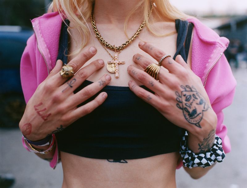 Close-up of a person wearing a pink hoodie, black crop top, layered gold chains, and multiple rings and hand tattoos.