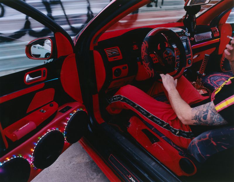 Driver in red sweatpants sits inside a heavily customized red and black car interior with speakers on the open door.