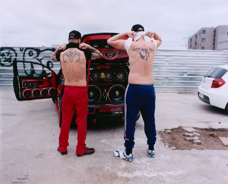 Two shirtless men with back tattoos standing with their backs to the camera in front of a red car with custom audio speakers.