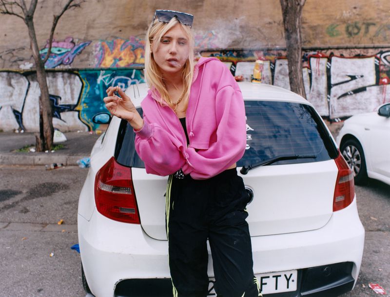 Blonde woman in a pink zip-up hoodie leaning against a white hatchback car in front of a graffiti wall
