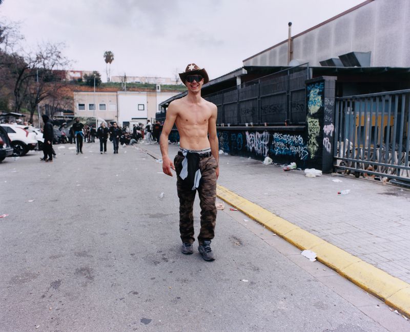Shirtless young man wearing a cowboy hat and sunglasses walking on an outdoor asphalt street for Dazed Magazine.