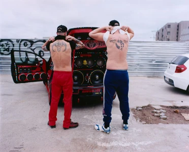 Two shirtless men with back tattoos standing with their backs to the camera in front of a red car with custom audio speakers.