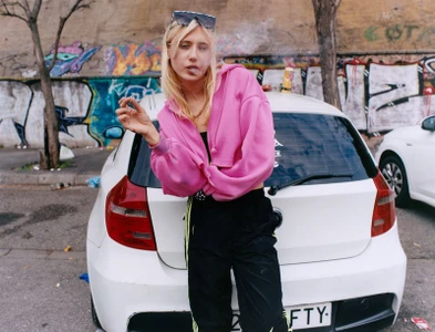 Blonde woman in a pink zip-up hoodie leaning against a white hatchback car in front of a graffiti wall