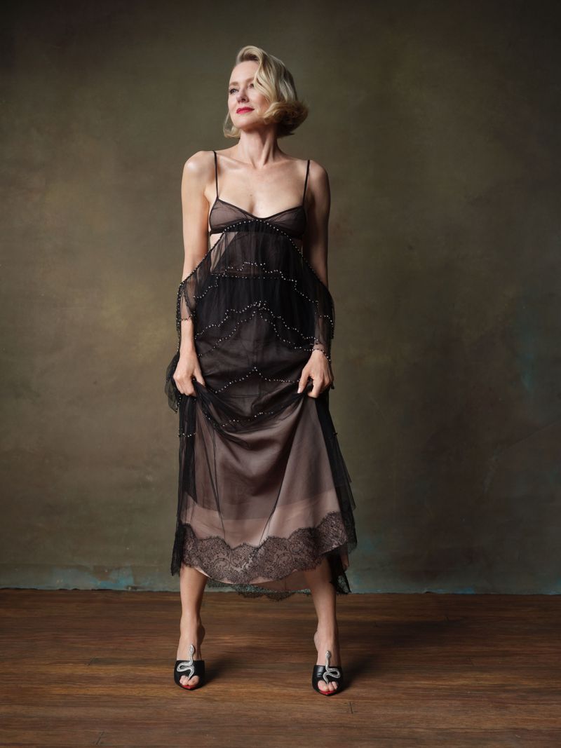 A fair-skinned woman in a black dress with black high-heeled shoes stands against a textured background in a full-body portrait.