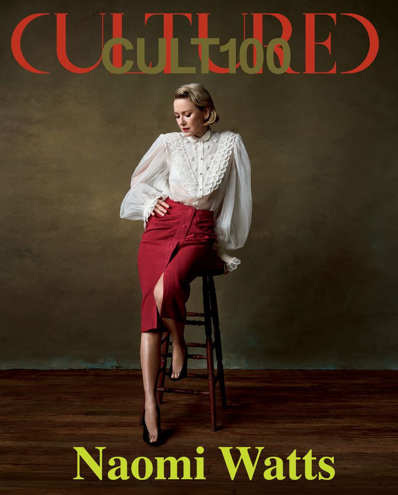Naomi Watts seated on a wooden stool wearing a white ruffled blouse and red skirt against a moody dark backdrop for Cultured Magazine.