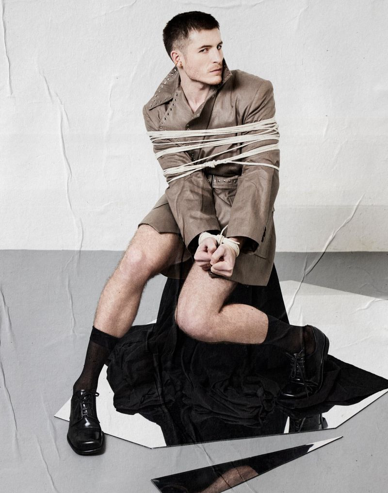 Man seated wearing a beige coat bound in rope with hands tied, over a mirror surface against a textured background.