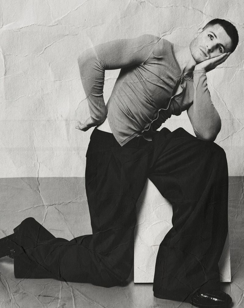 Adult male in long-sleeve shirt and loose pants poses while kneeling on a white block in a studio with a black-and-white filter.