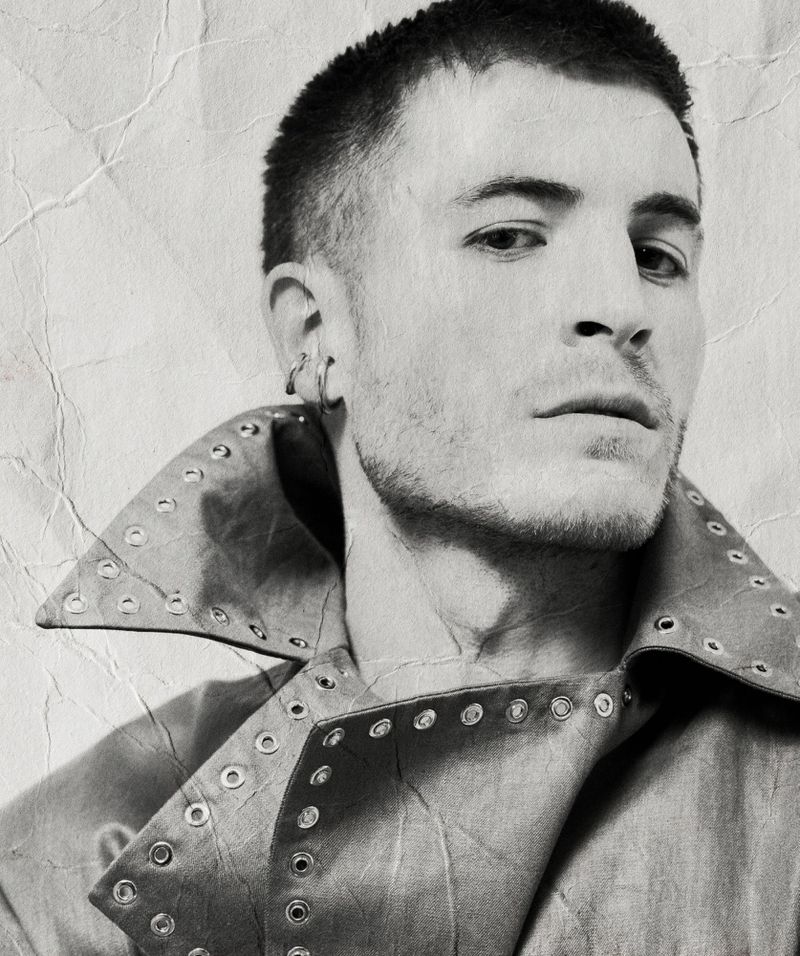 Portrait of a man wearing a studded jacket with short hair and ear cuffs against a distressed background.