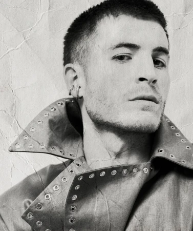 Portrait of a man wearing a studded jacket with short hair and ear cuffs against a distressed background.