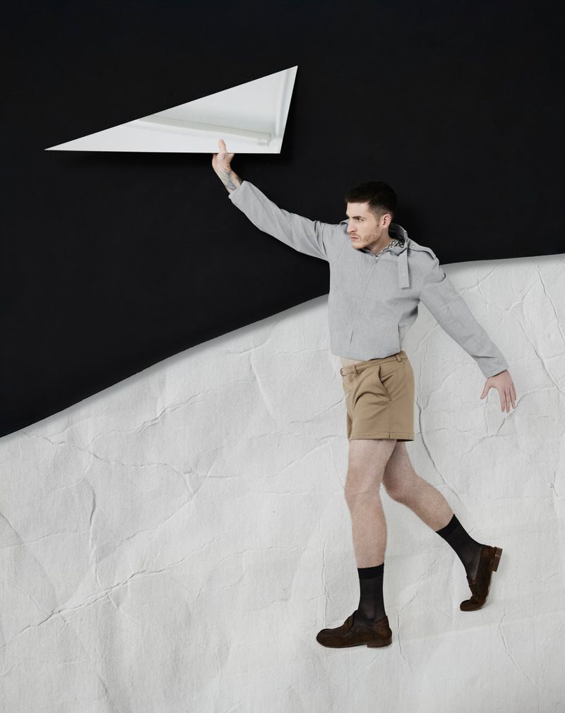 Man wearing grey jacket and khaki shorts holding a white geometric prop against a textured backdrop