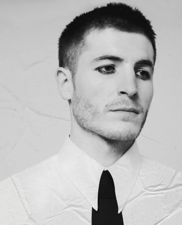Portrait of a man with short hair and dark eye makeup wearing a white collared shirt and black tie against a light background