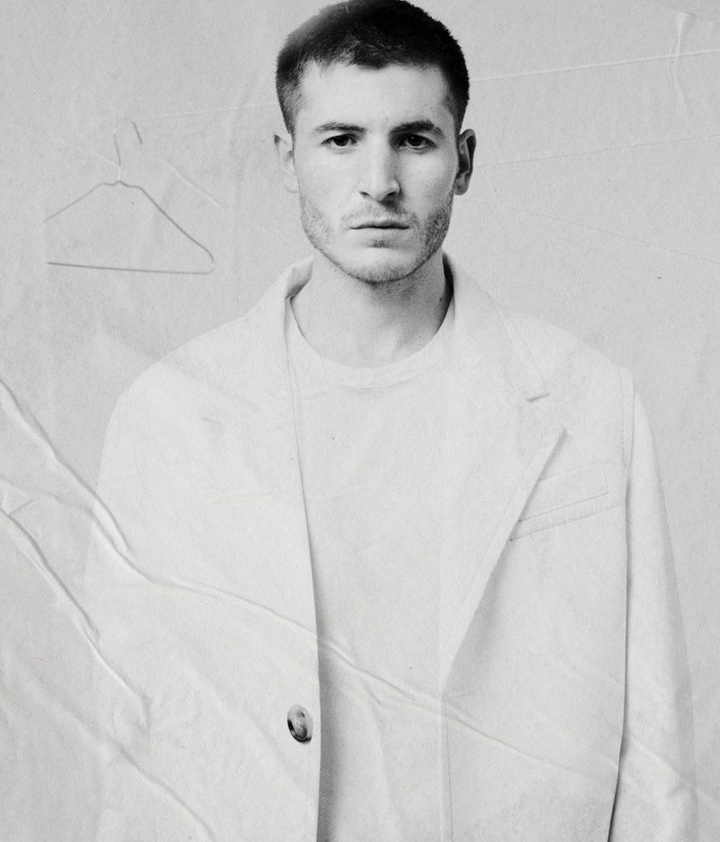 Portrait of a man wearing a light blazer and t-shirt against a textured, creased backdrop with a faint hanger outline