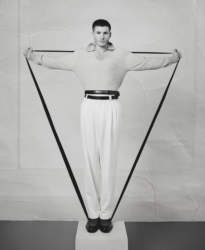 Monochrome portrait of a man holding a black strap forming a triangle shape, wearing light sweater, white trousers, and dark loafers.