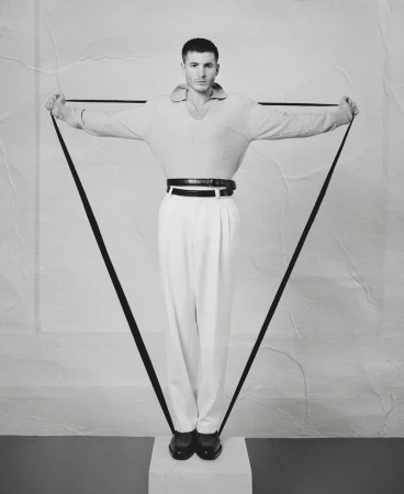 Monochrome portrait of a man holding a black strap forming a triangle shape, wearing light sweater, white trousers, and dark loafers.