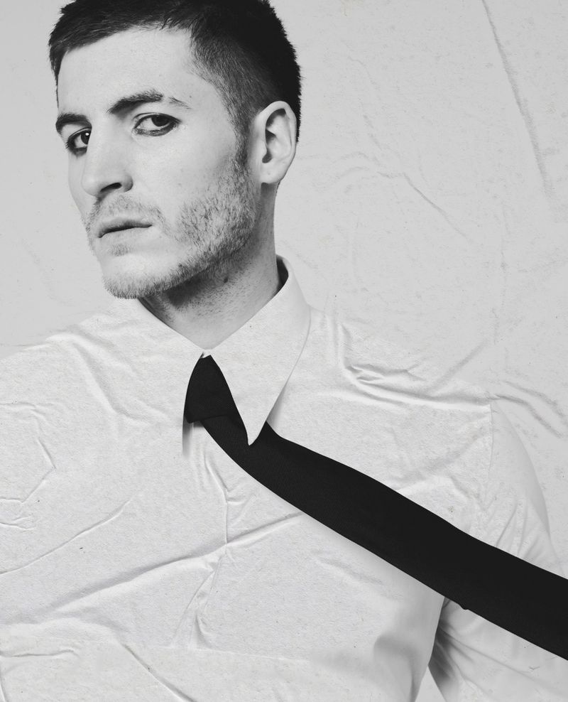 Portrait of a man with dark eye makeup wearing a white shirt and black tie against a textured backdrop for Le Mile Magazine.
