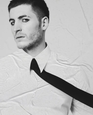 Portrait of a man with dark eye makeup wearing a white shirt and black tie against a textured backdrop for Le Mile Magazine.
