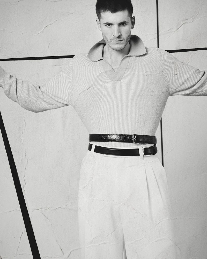 Man wearing a textured polo shirt and trousers with two belts against a geometric wall in a black and white editorial portrait.