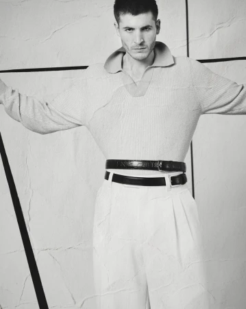 Man wearing a textured polo shirt and trousers with two belts against a geometric wall in a black and white editorial portrait.