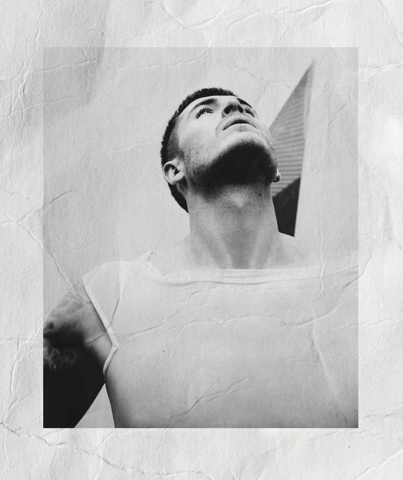 Monochrome portrait of a man looking upward wearing a white t-shirt against a textured, creased background.