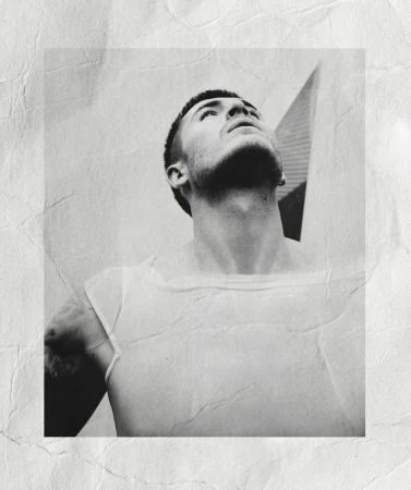 Monochrome portrait of a man looking upward wearing a white t-shirt against a textured, creased background.