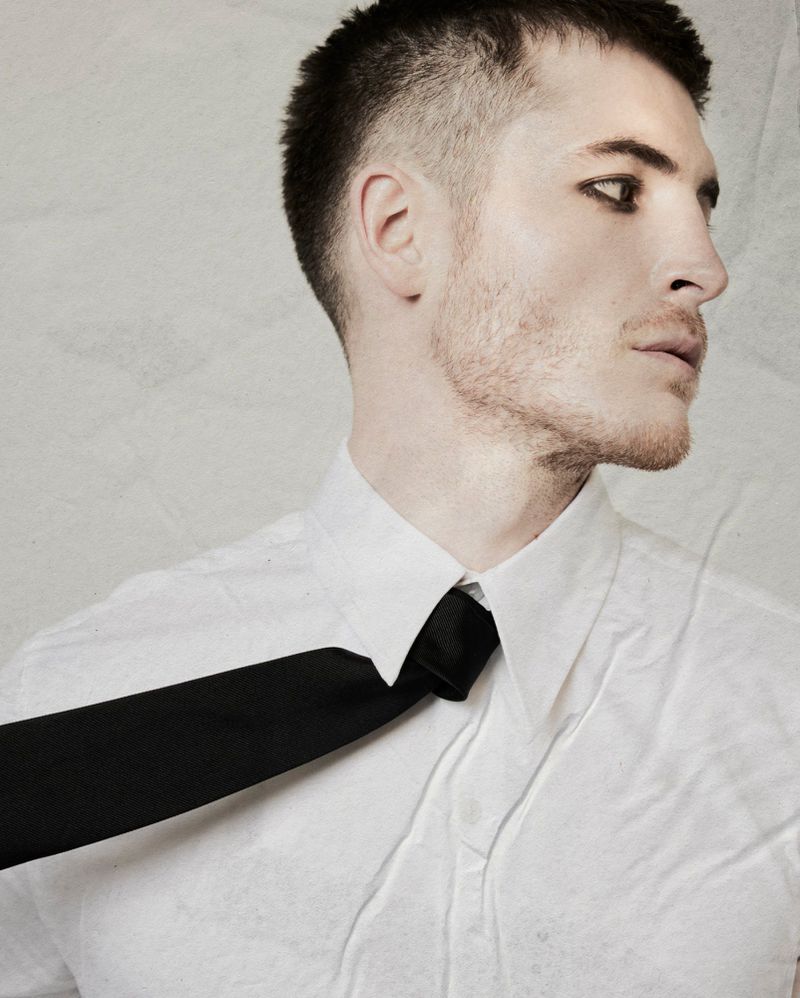 Close-up portrait of a man in a white shirt and black tie against a textured neutral background for Le Mile Magazine.