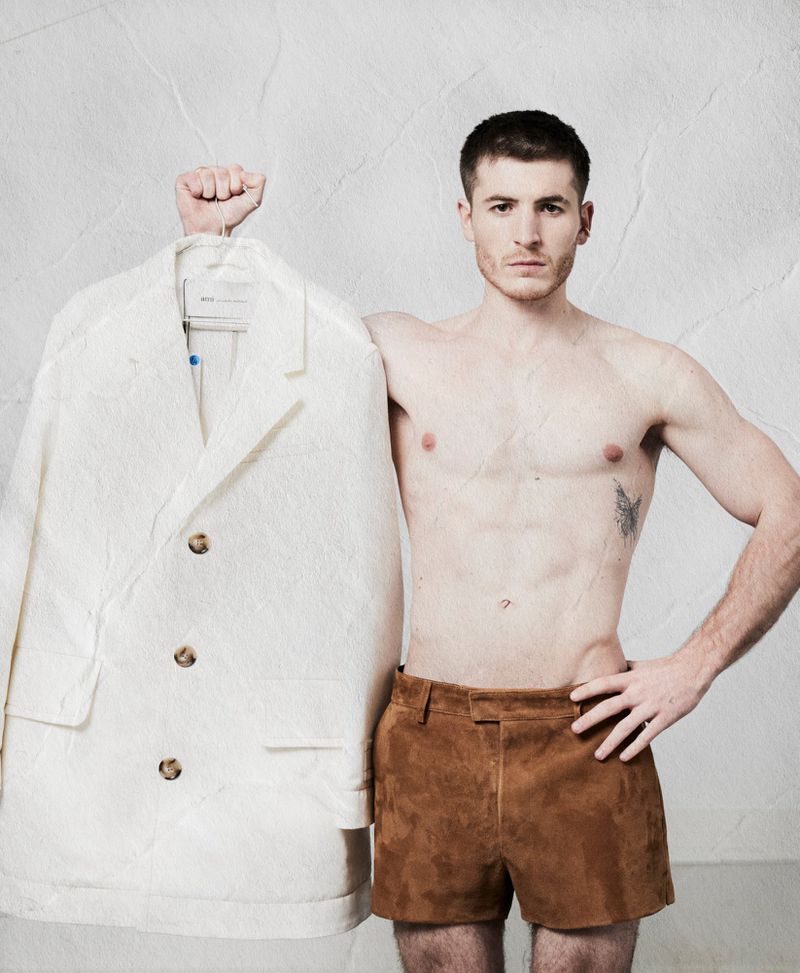 Shirtless man wearing brown shorts holding up a white textured coat on a hanger against a textured wall