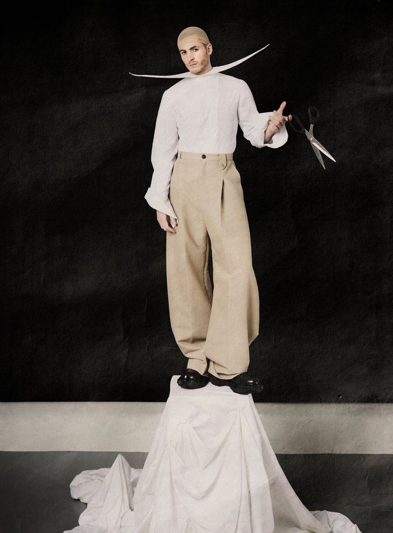 Man wearing a white shirt with an extended collar and beige trousers holding scissors while standing on a draped pedestal.