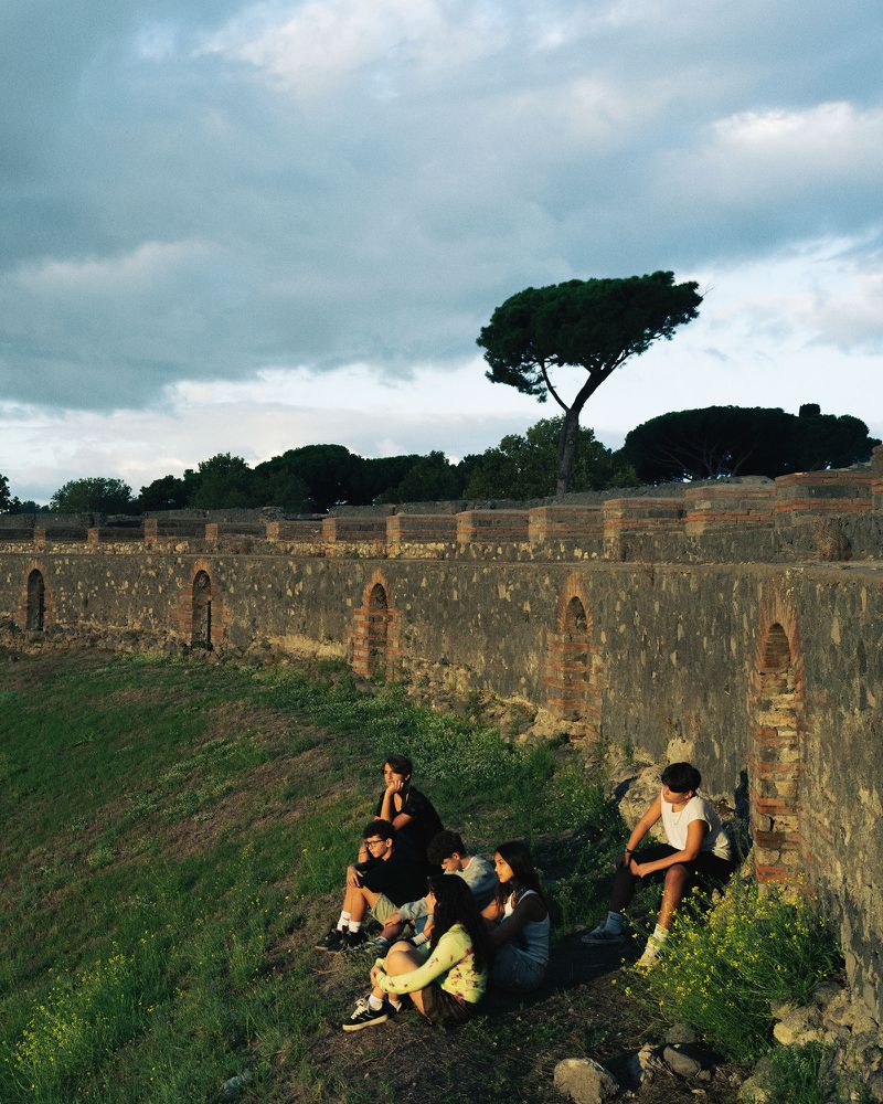 Group of young people seated together on a grassy slope below an ancient stone wall beneath a cloudy sky.