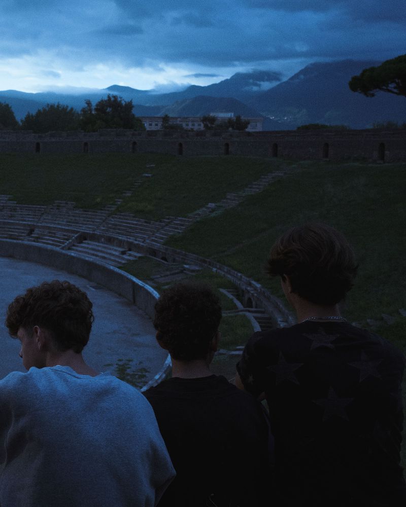 Three young men viewed from behind overlooking an ancient stone amphitheater under a dark twilight sky.