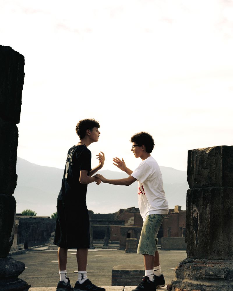 Two teenage boys facing each other outdoors among ancient stone ruins with a pale sky and distant mountains in the background