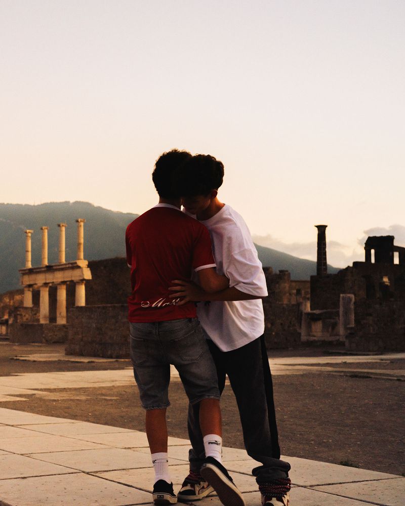 Two young men embracing amid ancient Roman ruins at dusk with distant mountains under a soft sky