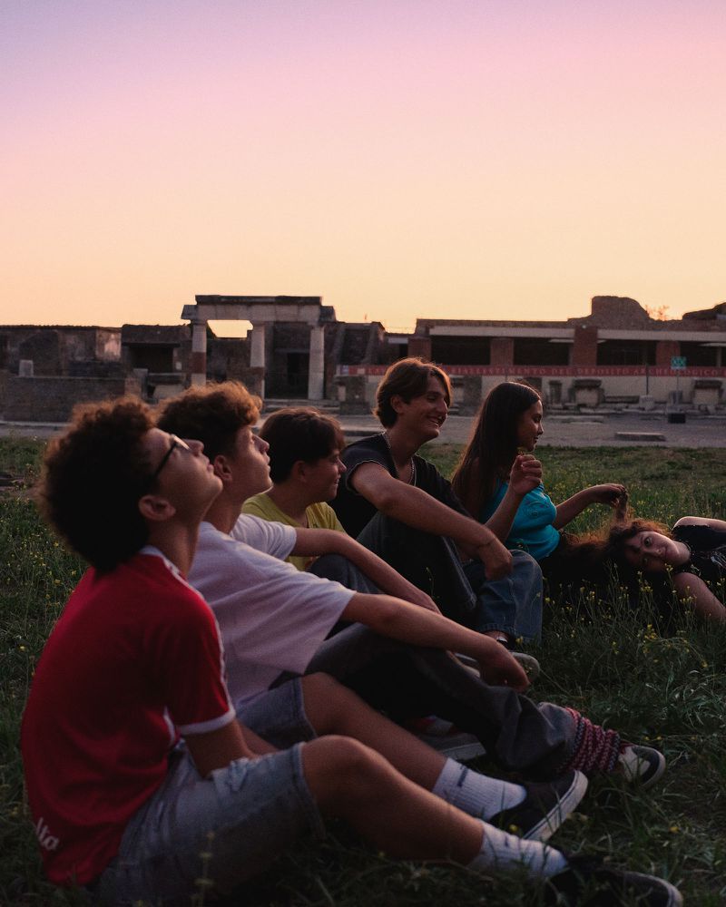 Group of young people sitting on grassy ground looking up toward ancient ruins under a warm sunset sky.