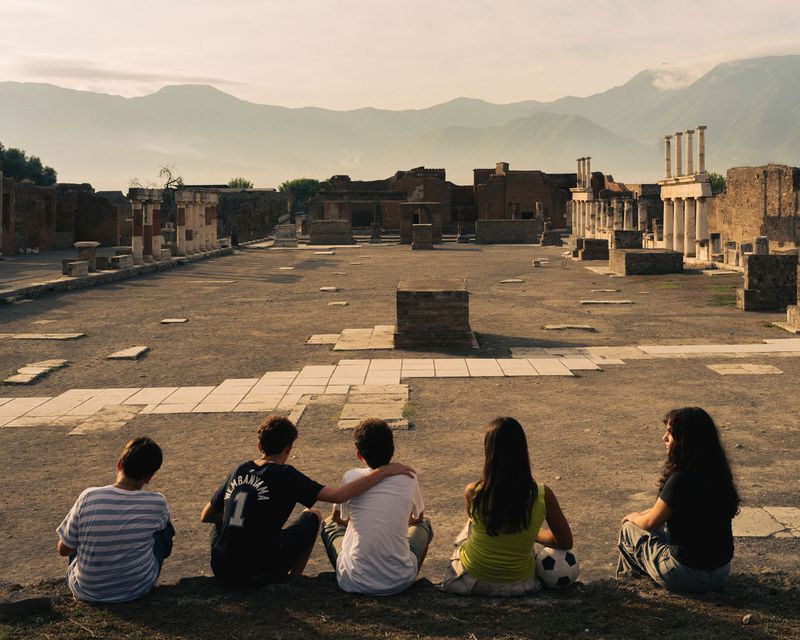 Five youths sitting in a row viewed from behind, looking out over ancient ruins beneath distant hazy mountains.