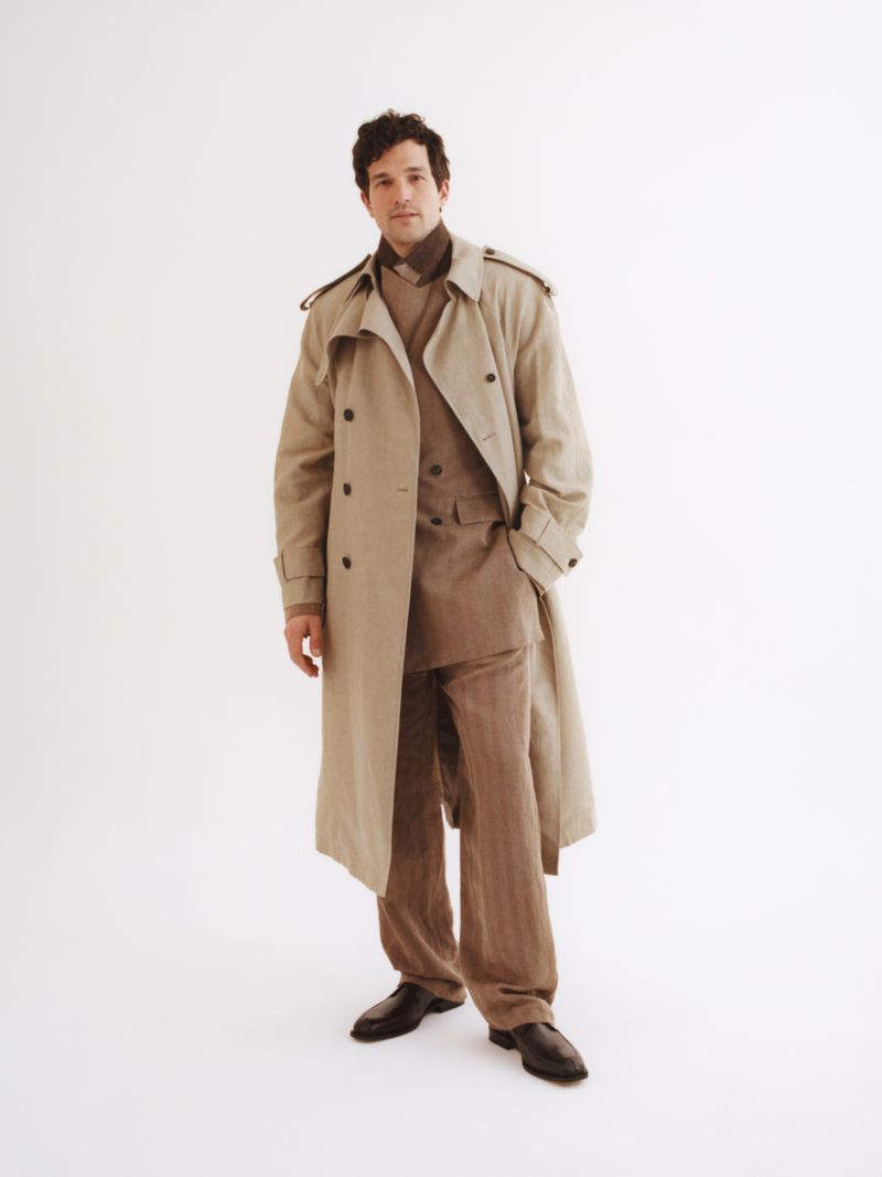 Man wearing a beige trench coat over a brown suit standing against a plain white background