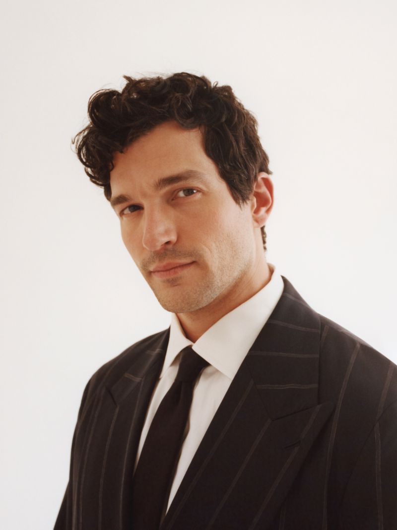 Portrait of an adult male with dark curly hair wearing a pinstripe suit, white collared shirt and black tie against a white backdrop.