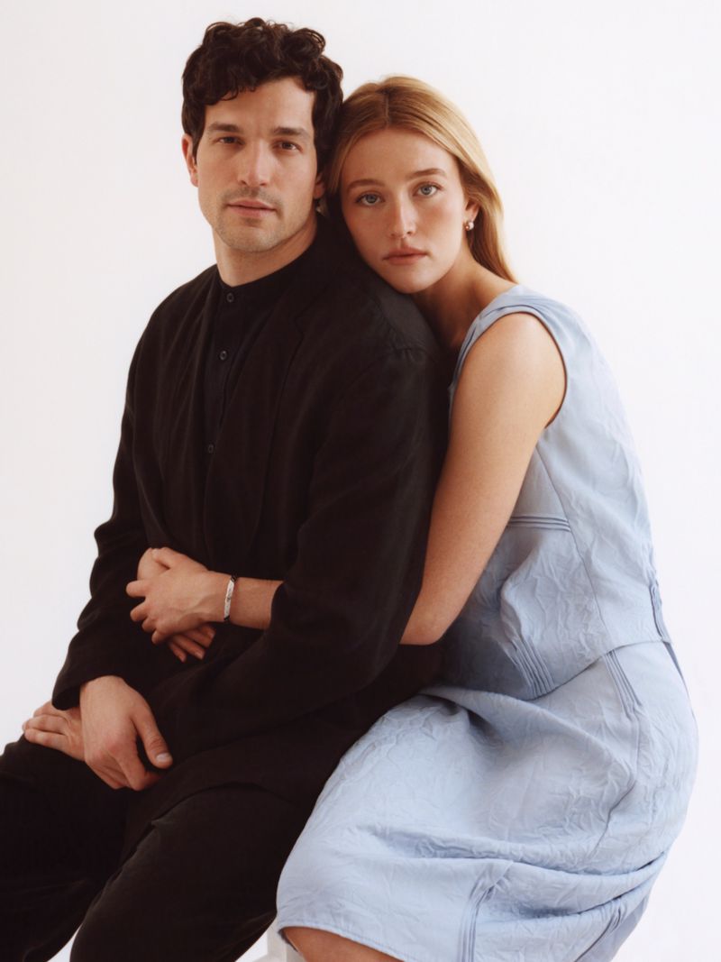 Portrait of a man in a black shirt sitting with a woman in a light blue dress leaning on his shoulder against a plain white background