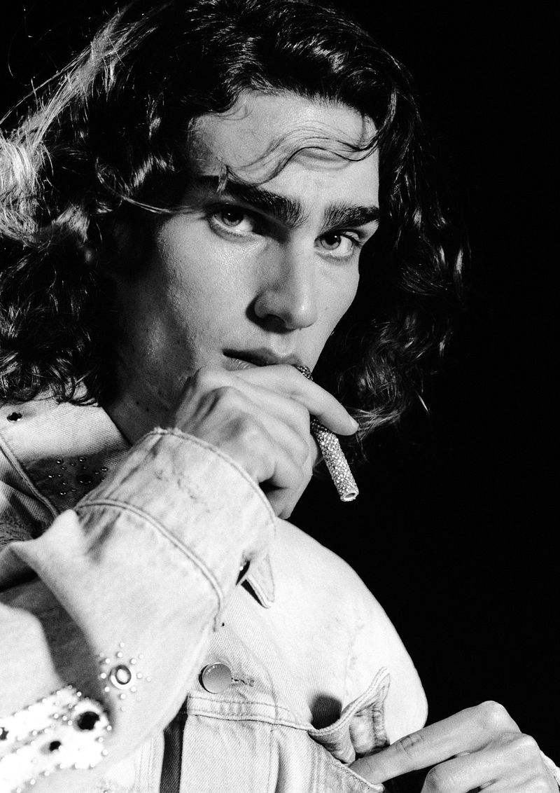 Black and white portrait of a long-haired man holding an embellished lighter to his mouth against a dark background
