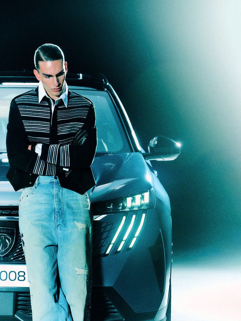 Male model in striped cardigan and light blue jeans leaning against a car in a studio setting for GQ Turkey editorial.