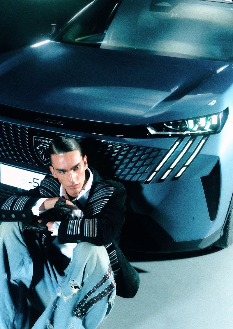 A seated model wearing a black patterned jacket and light jeans in front of a modern blue car for a GQ Turkey editorial.