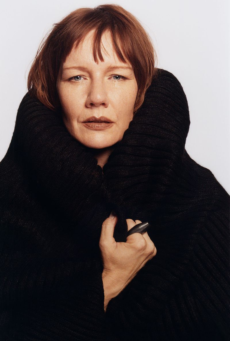 Portrait of a woman with auburn hair wearing a black knitted sweater with a large collar, posing against a light grey background.
