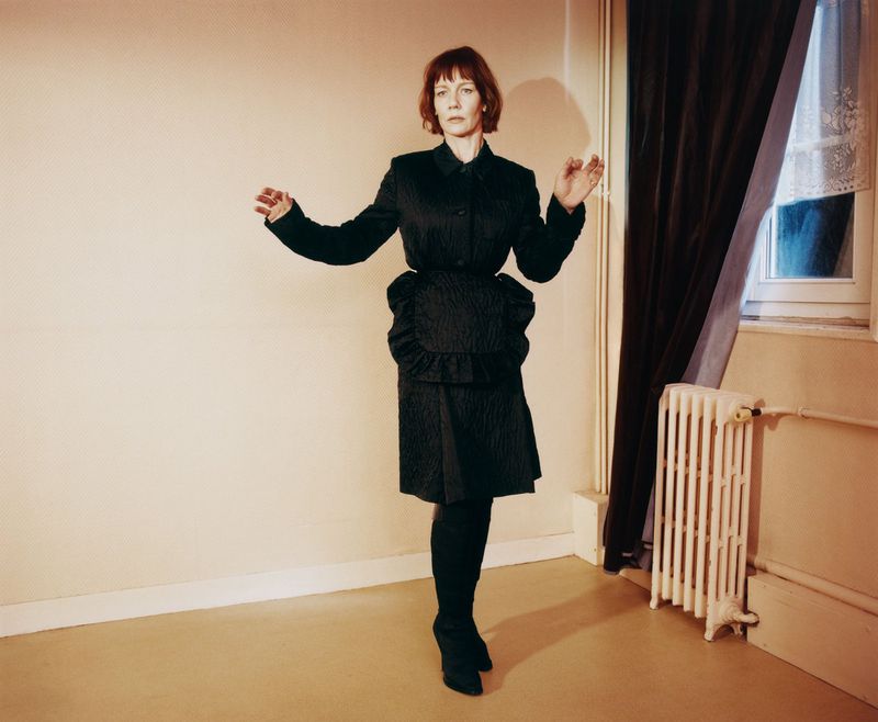 Sandra Hüller wearing a dark textured jacket and skirt set in a domestic room with a radiator and window.
