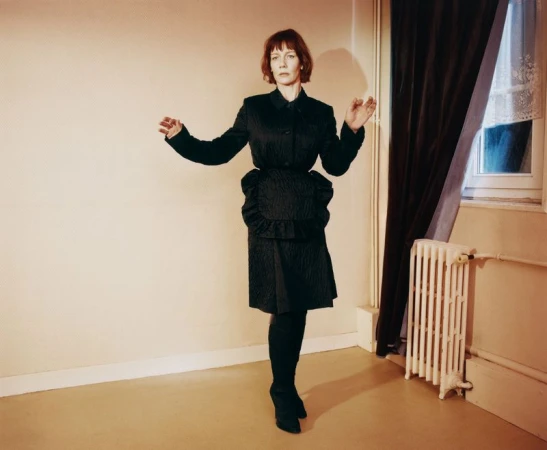 Sandra Hüller wearing a dark textured jacket and skirt set in a domestic room with a radiator and window.