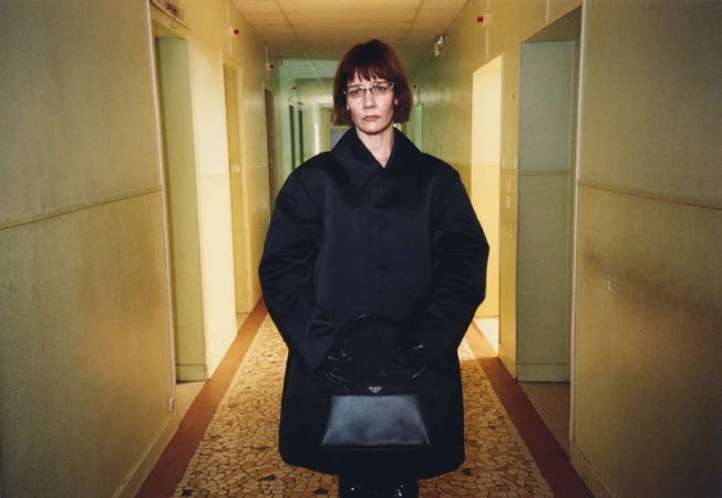 Sandra Huller in a black coat holding a handbag in a narrow hallway for In the Wings editorial by Family Style