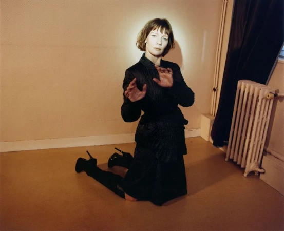 Sandra H\üller kneeling on a wooden floor in a black dress wearing high heels against a warm-toned interior wall.