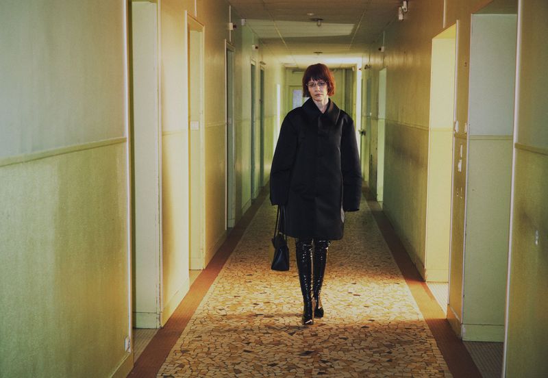 Sandra Huller in a black coat and tall boots walking down a long corridor for Family Style by Harley Weir