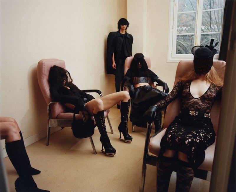 Group of models in dark clothing and veiled hats seated in pink armchairs within a neutral room.