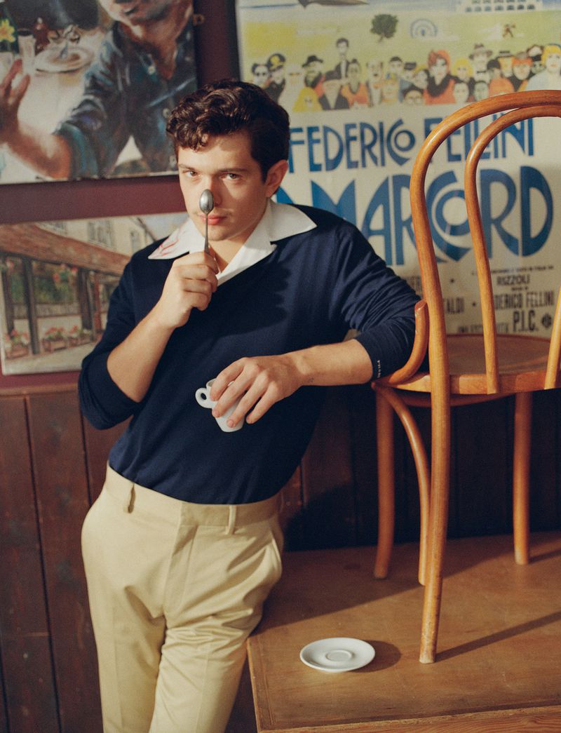 Noah Jupe holding a spoon to his nose and a small white cup, wearing a dark blue shirt and beige trousers in a vintage styled setting.