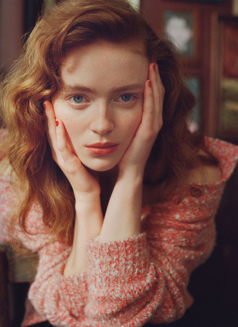 Portrait of Sadie Sink with hands on cheeks wearing a textured pink patterned sweater
